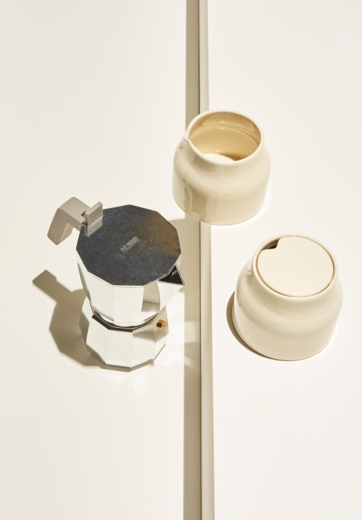 A silver octagonal coffee maker sits beside two cream ceramic cups and saucers against a white background