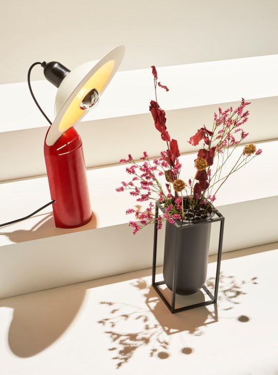 Red desk lamp and black vase with dried flowers on windowsill casting geometric shadows in natural light