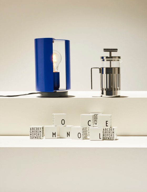 Blue cylindrical lamp with exposed bulb, silver French press, and wooden letter blocks arranged on white surface