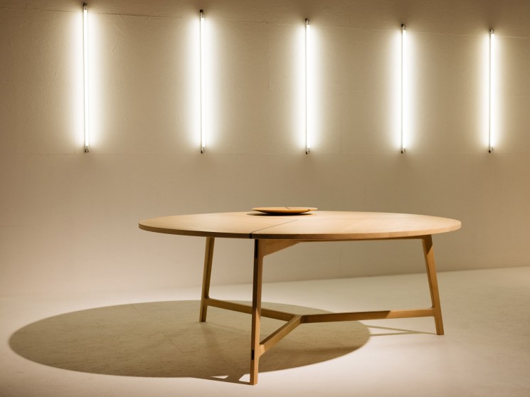 A minimalist oval wood dining table displayed against a warm wall with a row of vertical tube lights.