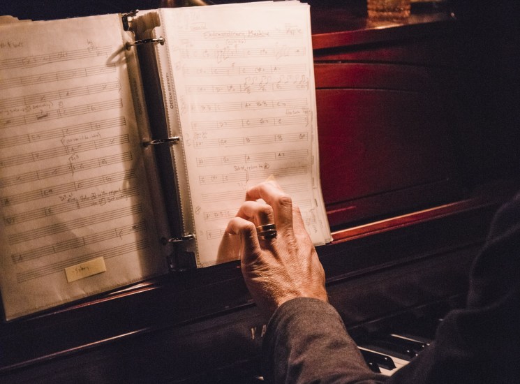 A pianist's hands turning sheet music pages at a candlelit upright piano in a dimly lit room.