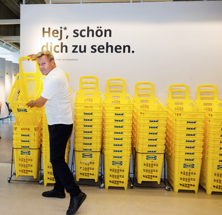 Man in white shirt pulling yellow IKEA shopping basket in store with stacks of yellow baskets and Swedish text on wall