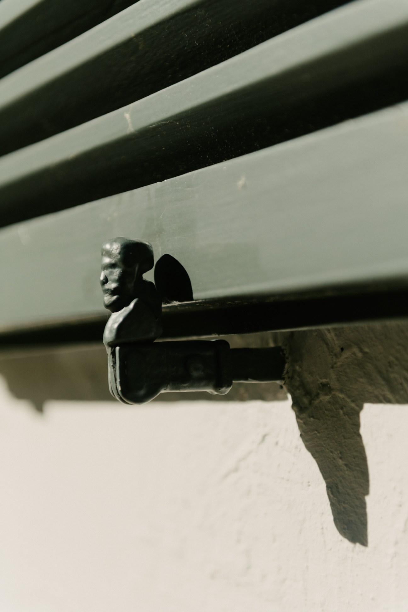 A silvered metal sculptural head mounted on a dark horizontal beam against a white and grey striped wall.