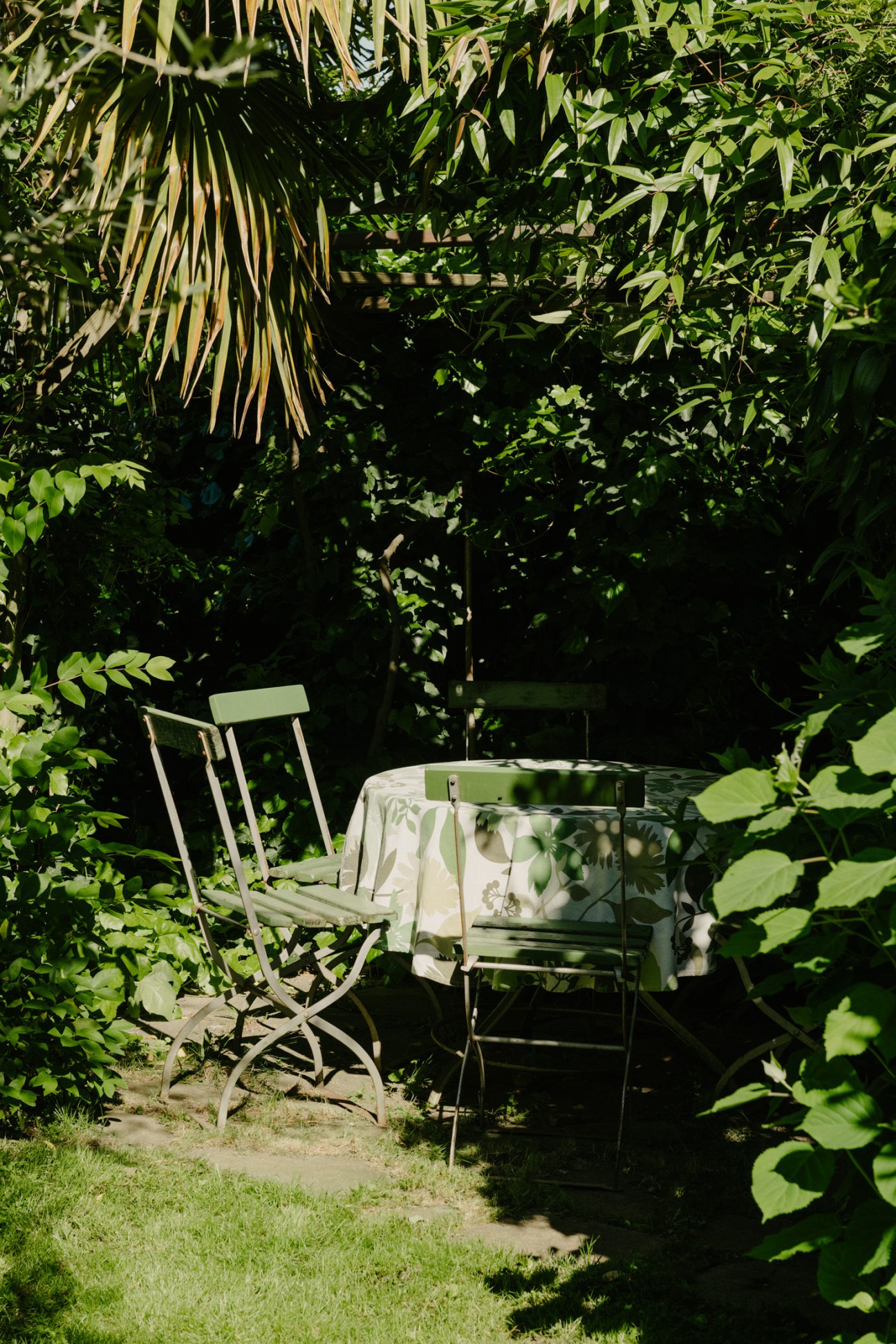Shaded outdoor dining area with metal café chairs and table surrounded by dense green foliage and vines.
