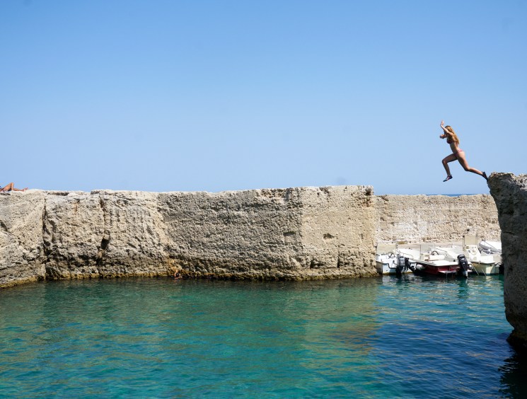 Person jumping from limestone cliffs into turquoise water with boats moored nearby