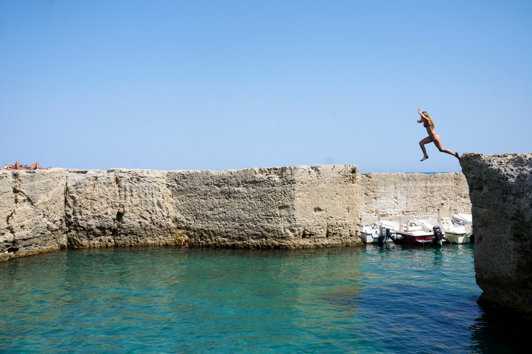 Person jumping from limestone cliffs into turquoise water with boats moored nearby