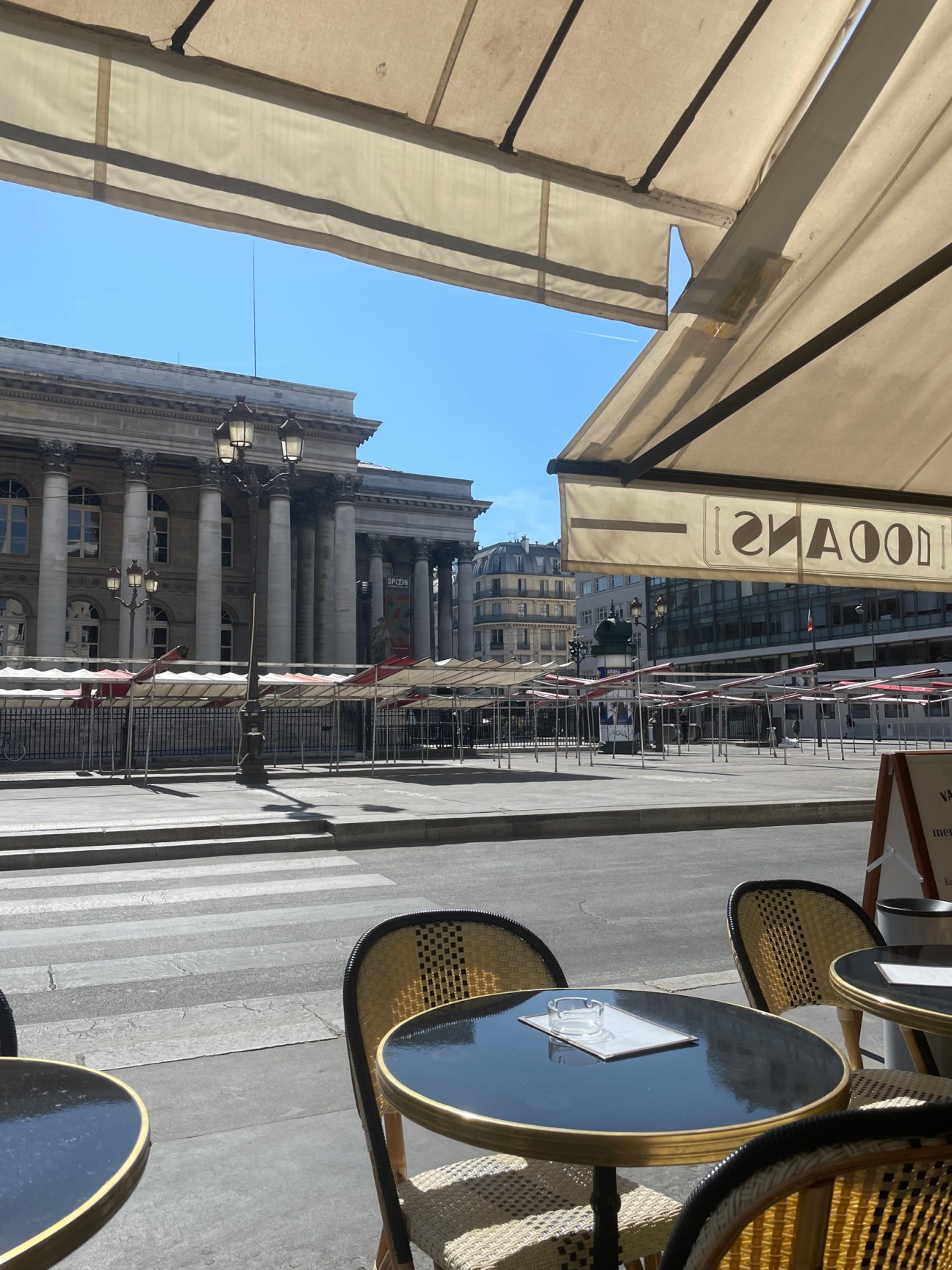 View from outdoor cafe terrace with tables facing a neoclassical building with columns across a paved square