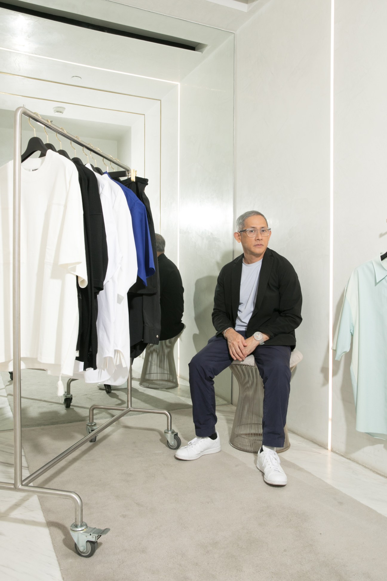Mark Gonzalez sitting on a wire basket chair in a white minimalist retail space with clothing racks