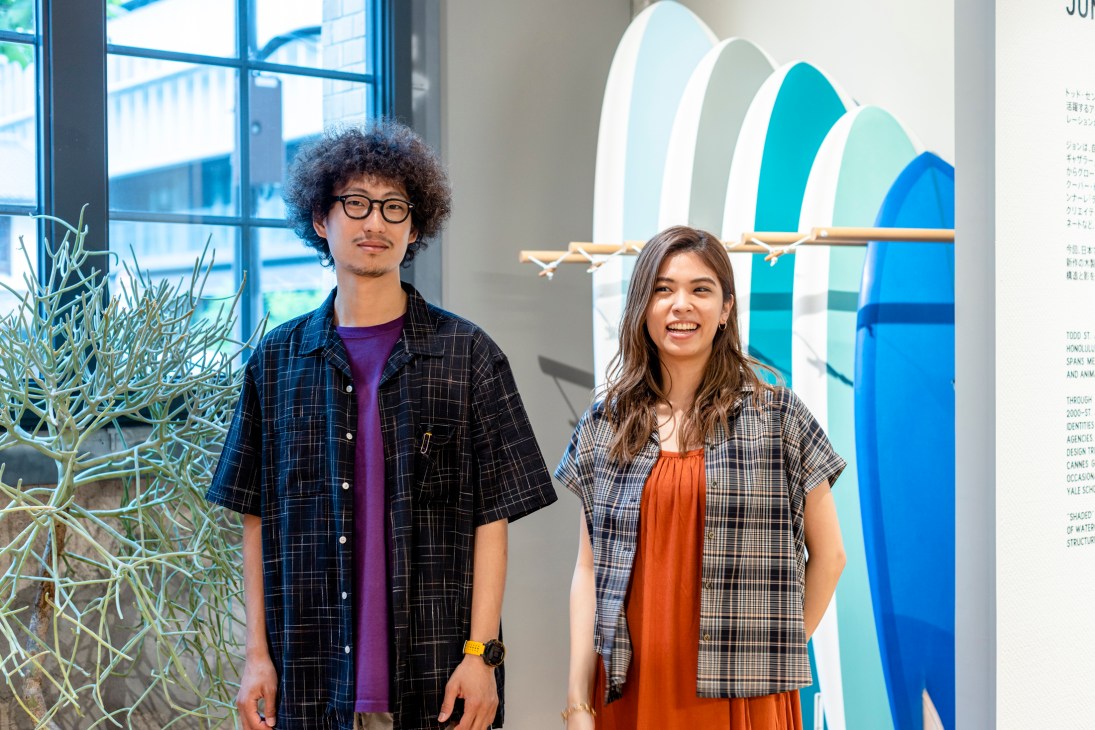 Two shop staff members stand in a bright retail space with colorful surfboard displays.