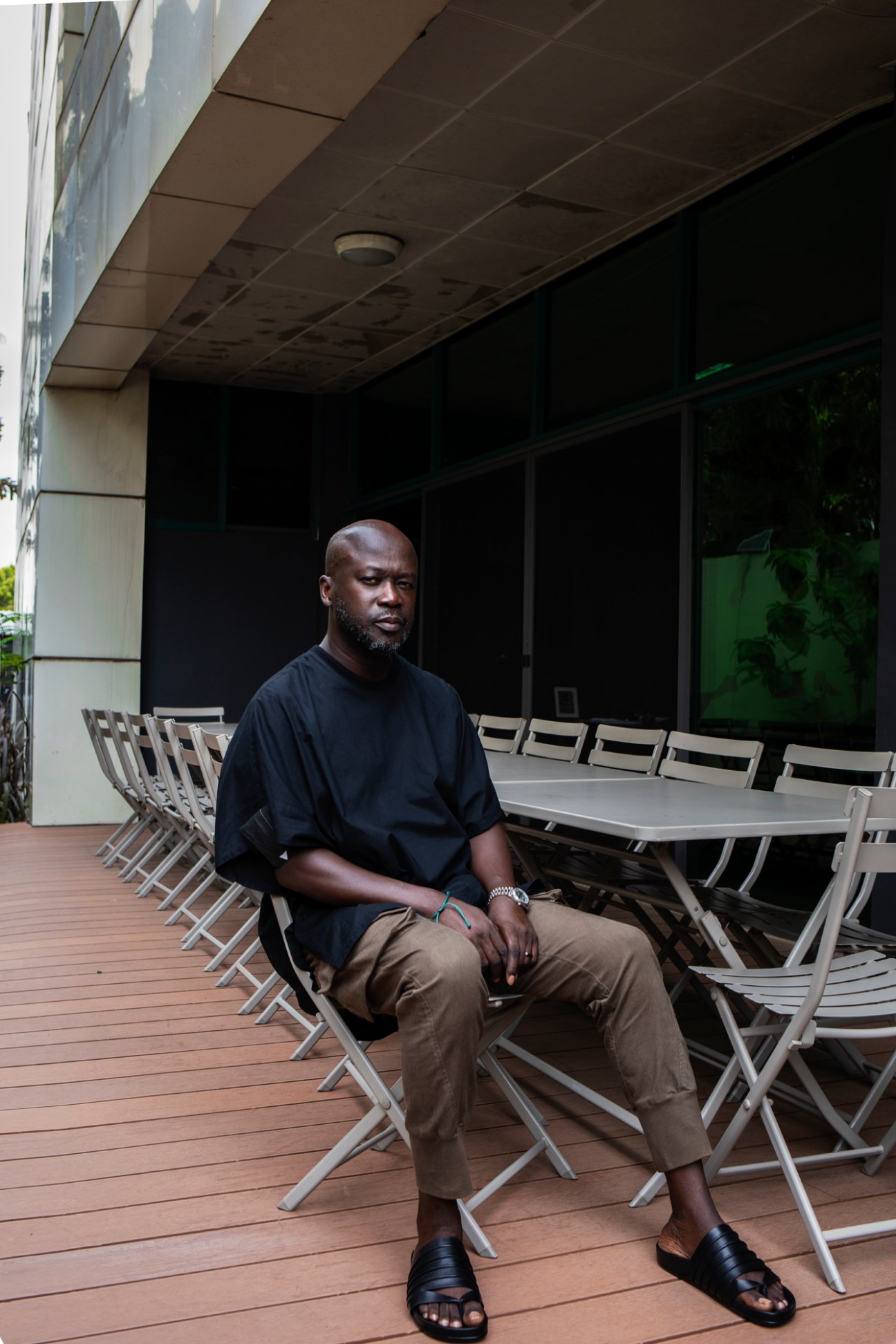 David Adjaye sits on a chair at an outdoor table on a wooden deck terrace