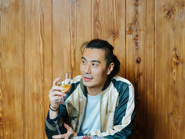A man holding a wine glass gazes to the side in front of a warm wood-panelled wall.
