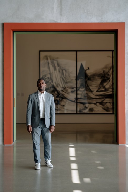 Franklin Sirmans stands in a modern art museum gallery with a red-framed entrance and sepia-toned artworks on display behind him.