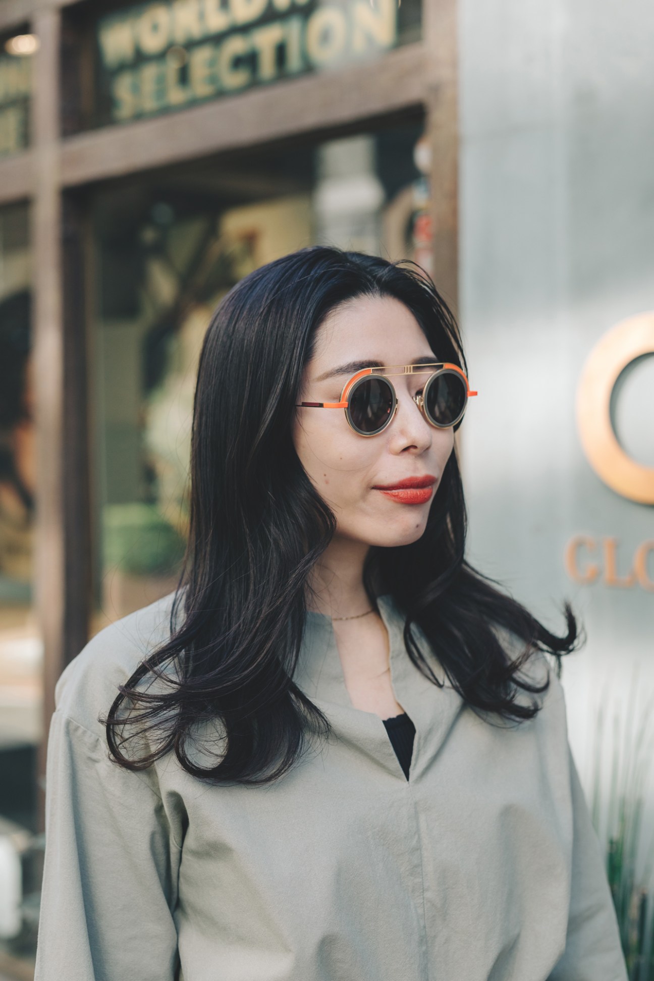 Woman wearing round sunglasses with orange and gold frames and dark lenses, styled in a light beige jacket over a black top, photographed outdoors in front of a blurred storefront.