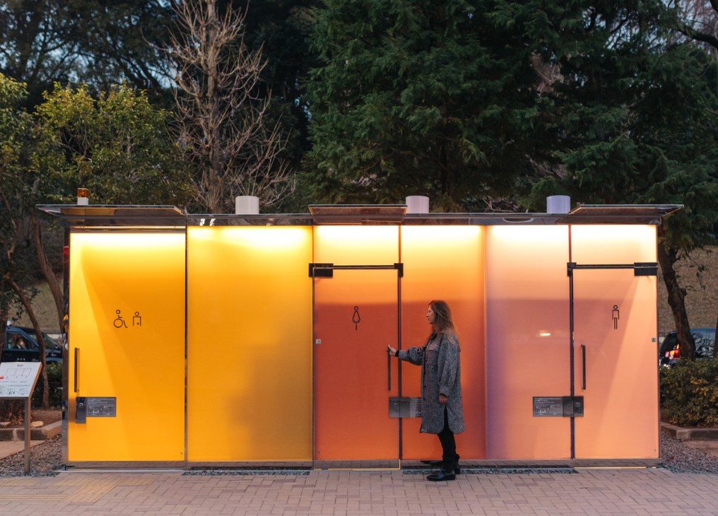 Woman standing by illuminated public toilet facility with translucent panels glowing orange and yellow at dusk