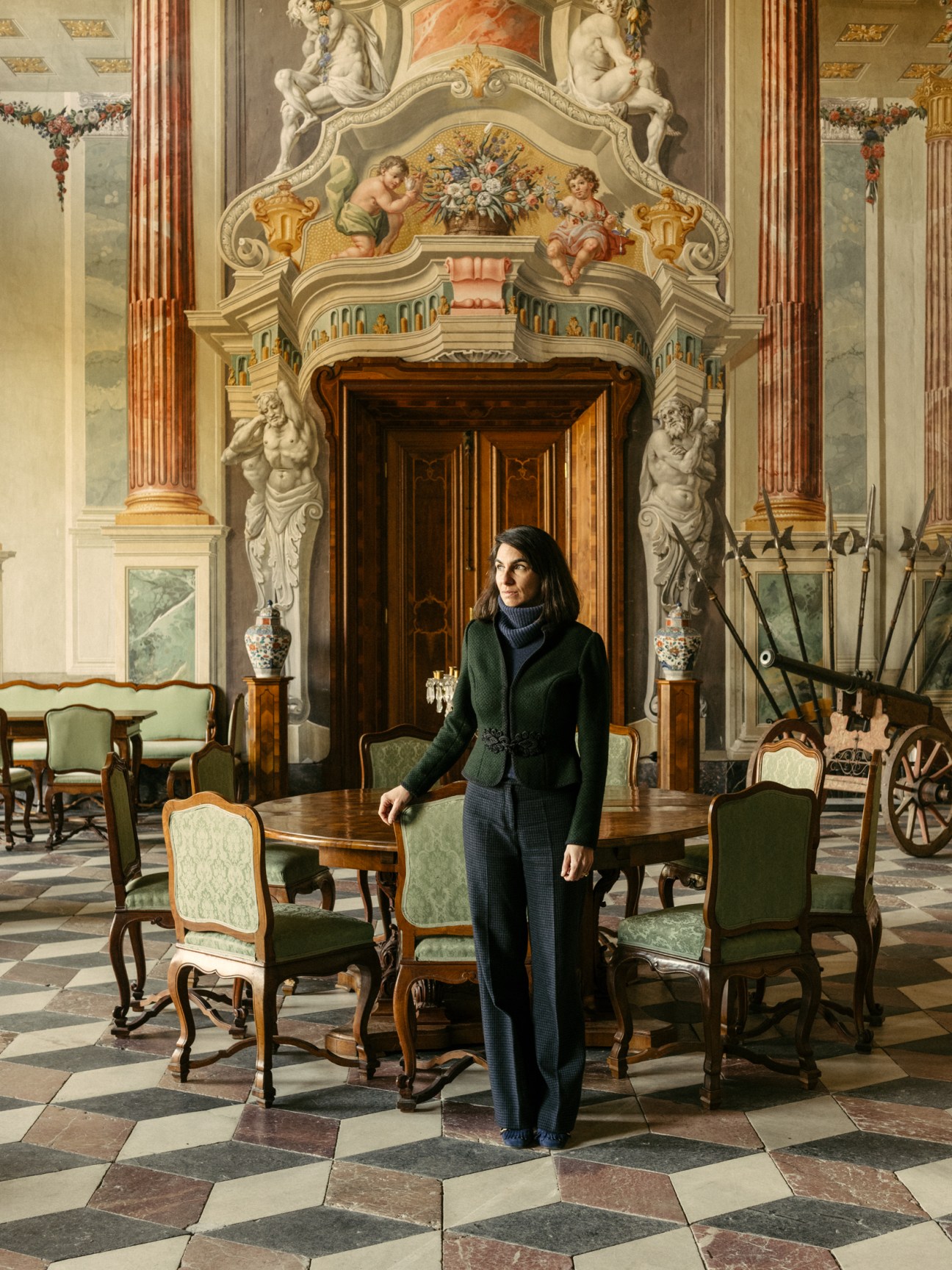 Alice Stori Liechtenstein standing beside a wooden table in an ornate baroque castle interior with decorated walls