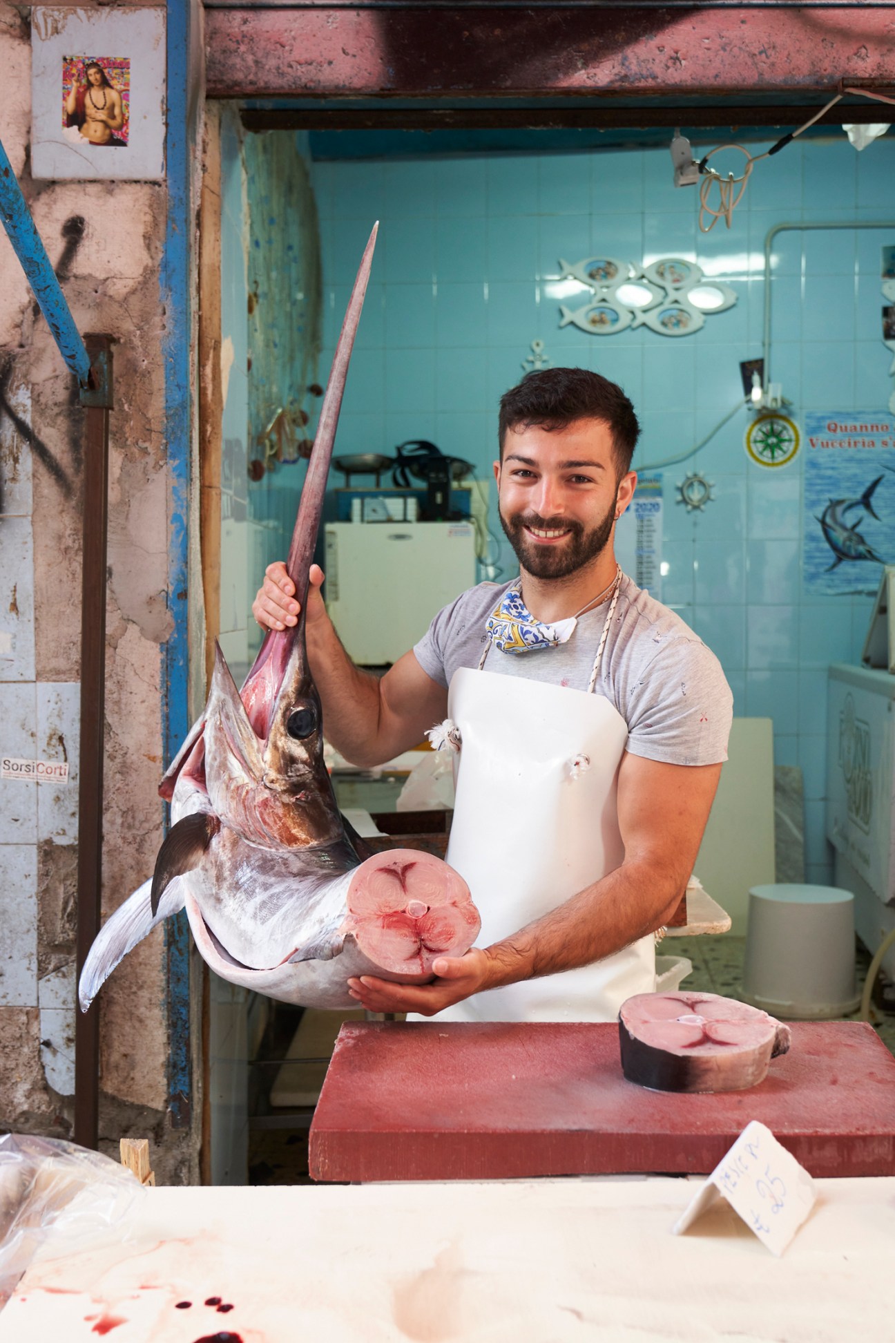 Fishmonger holding large swordfish at market stall with fish steaks displayed on cutting board