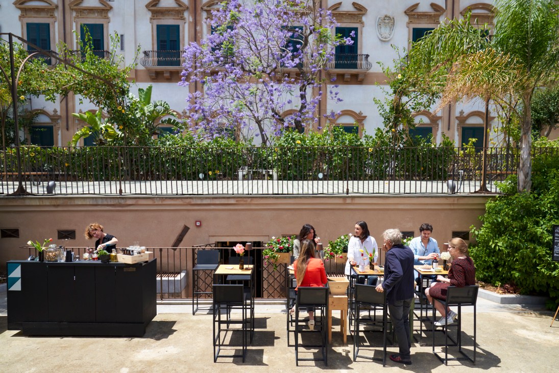 Outdoor restaurant terrace with diners seated at tables beneath blooming jacaranda tree and historic Sicilian palazzo