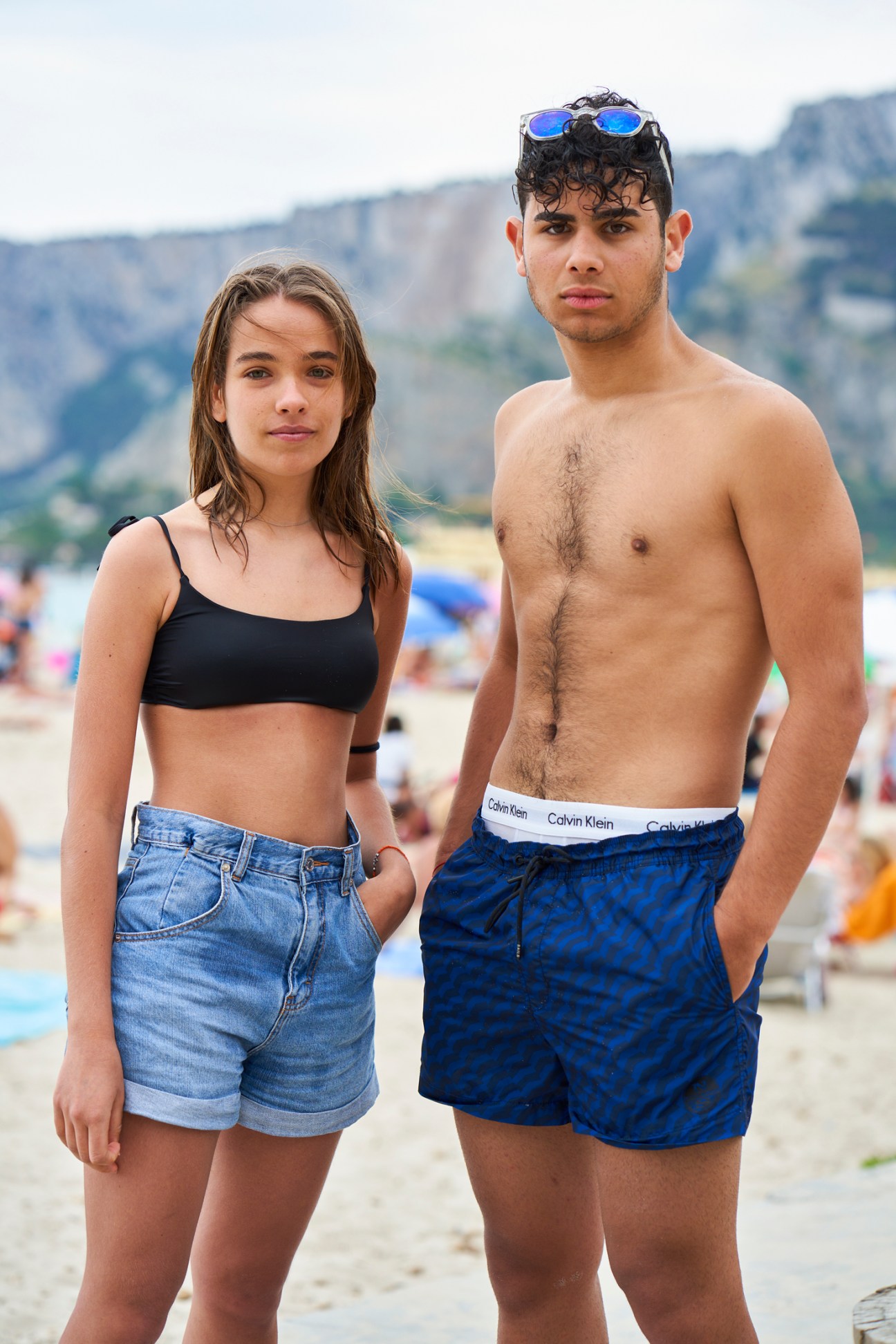 Young woman in black bikini top and denim shorts standing next to shirtless young man in blue swim shorts on beach