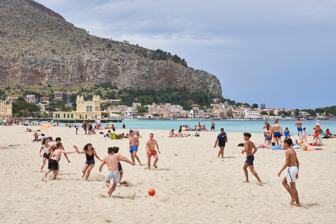 People playing soccer and relaxing on Mondello Beach with turquoise water and coastal buildings in background