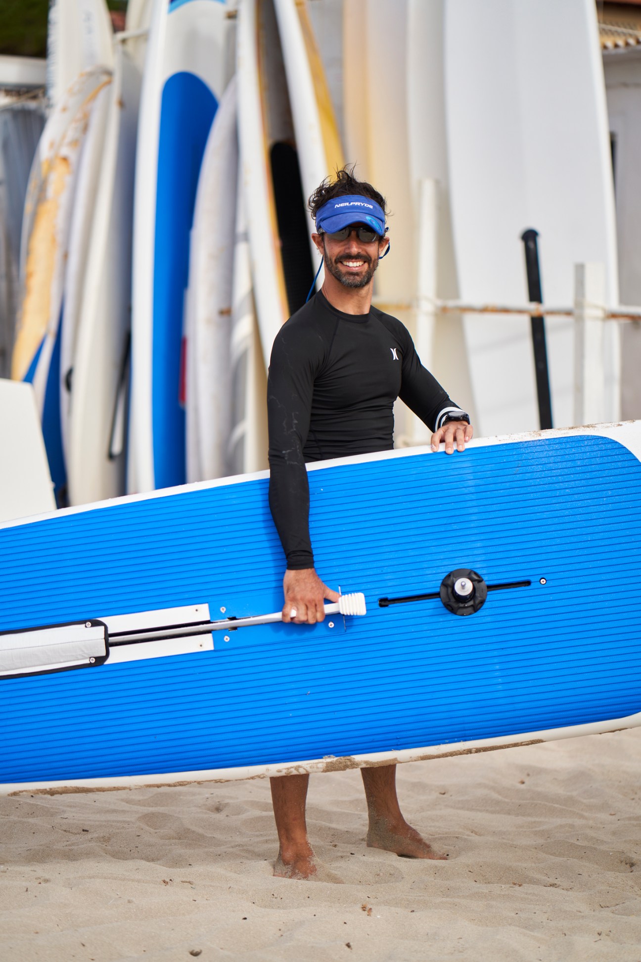 Windsurfer Guido Piacere holds a blue paddle board while standing on a beach near white surfboards