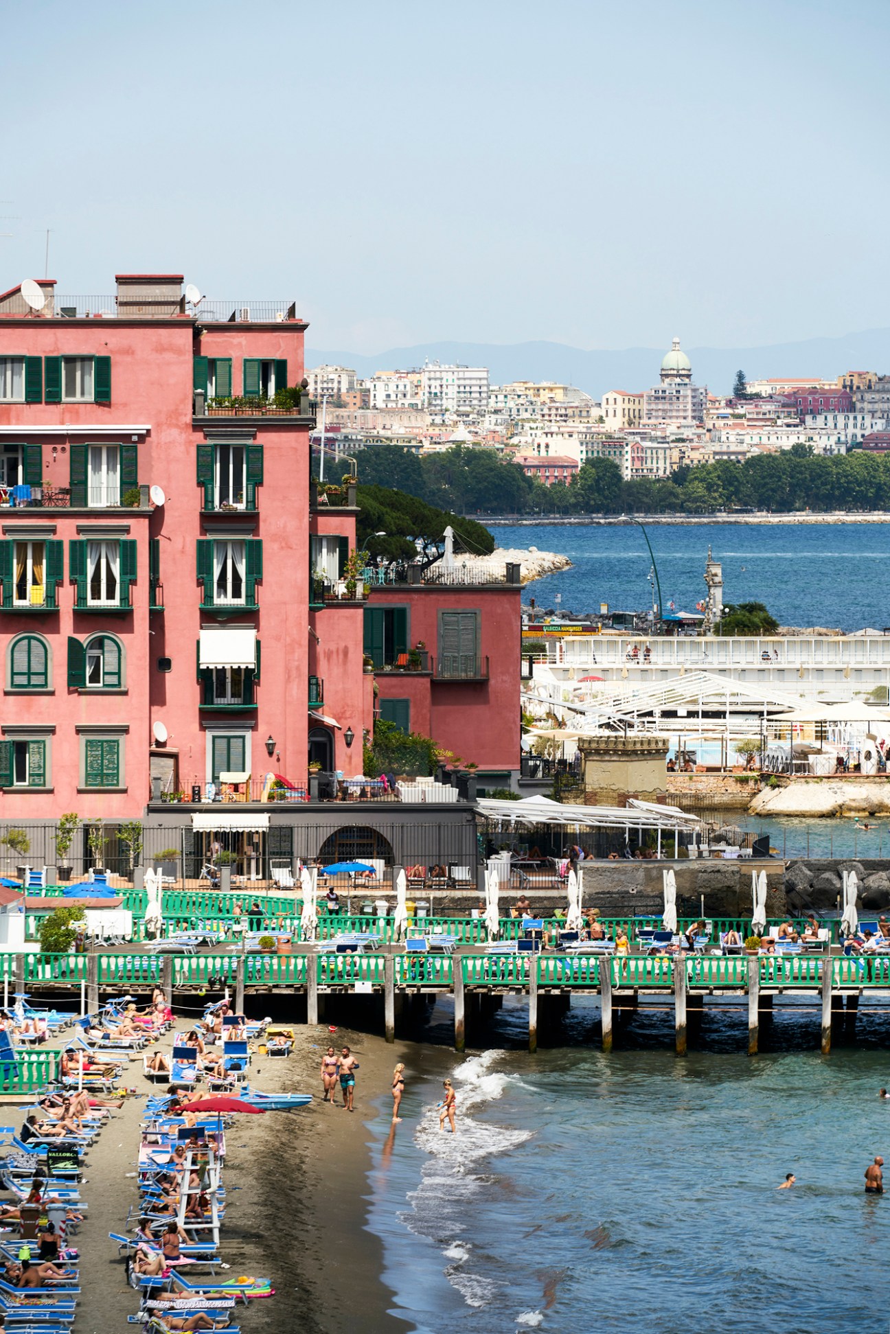 Coastal Italian town with pink buildings overlooking beach with wooden pier and beachgoers, Naples across the bay