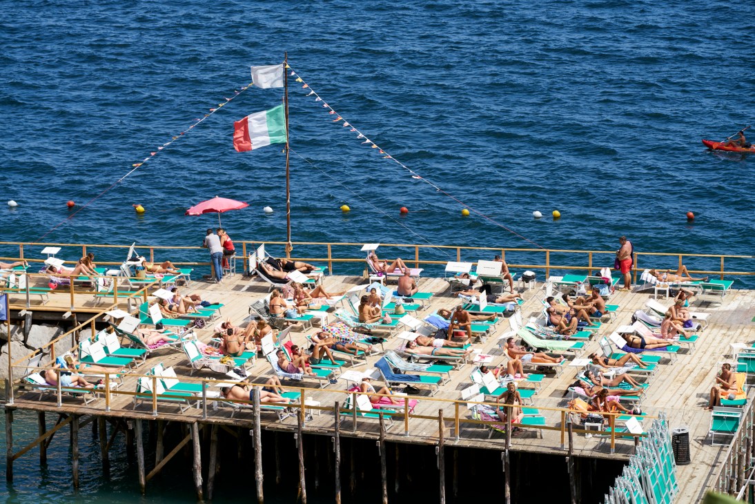 Crowded wooden pier at Bagno Sirena beach club with Italian flag flying, sunbathers on turquoise loungers overlooking blue sea