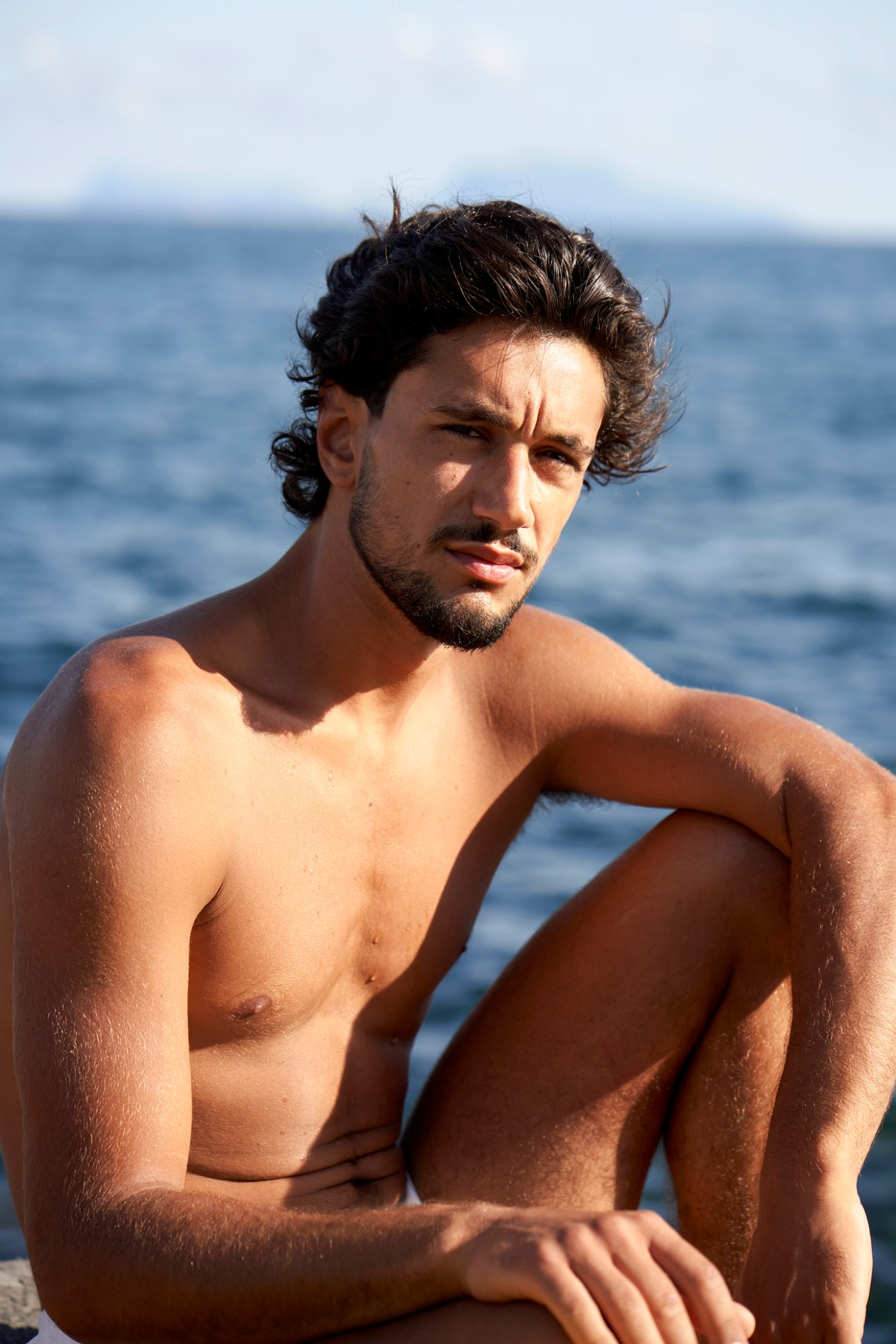 Shirtless man with curly dark hair and beard sitting by the ocean at Lido Marechiaro