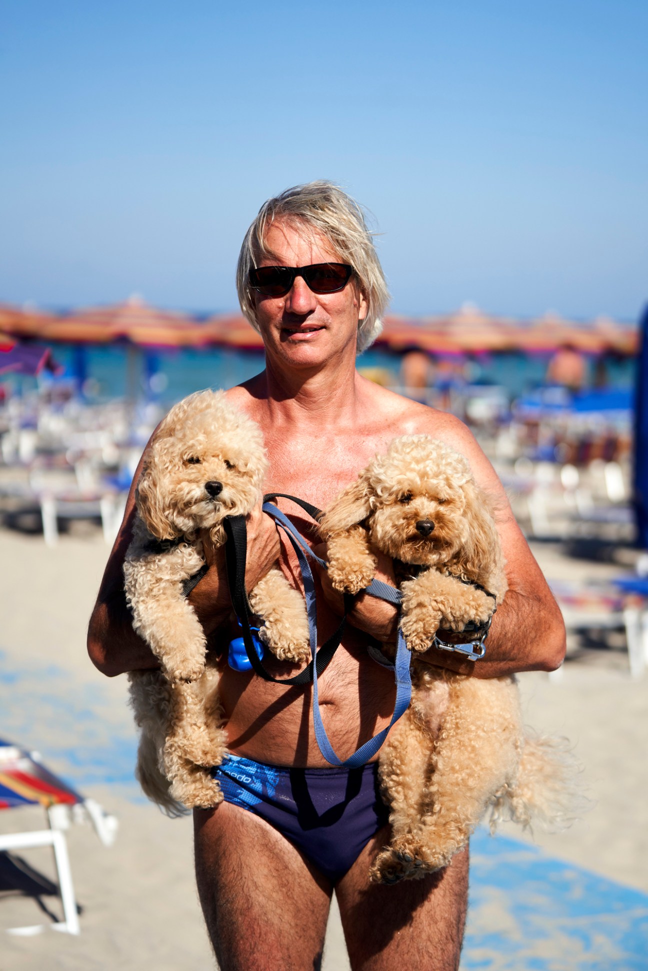 Blonde woman in swimwear holding two small poodle dogs with blue harnesses at an Italian beach