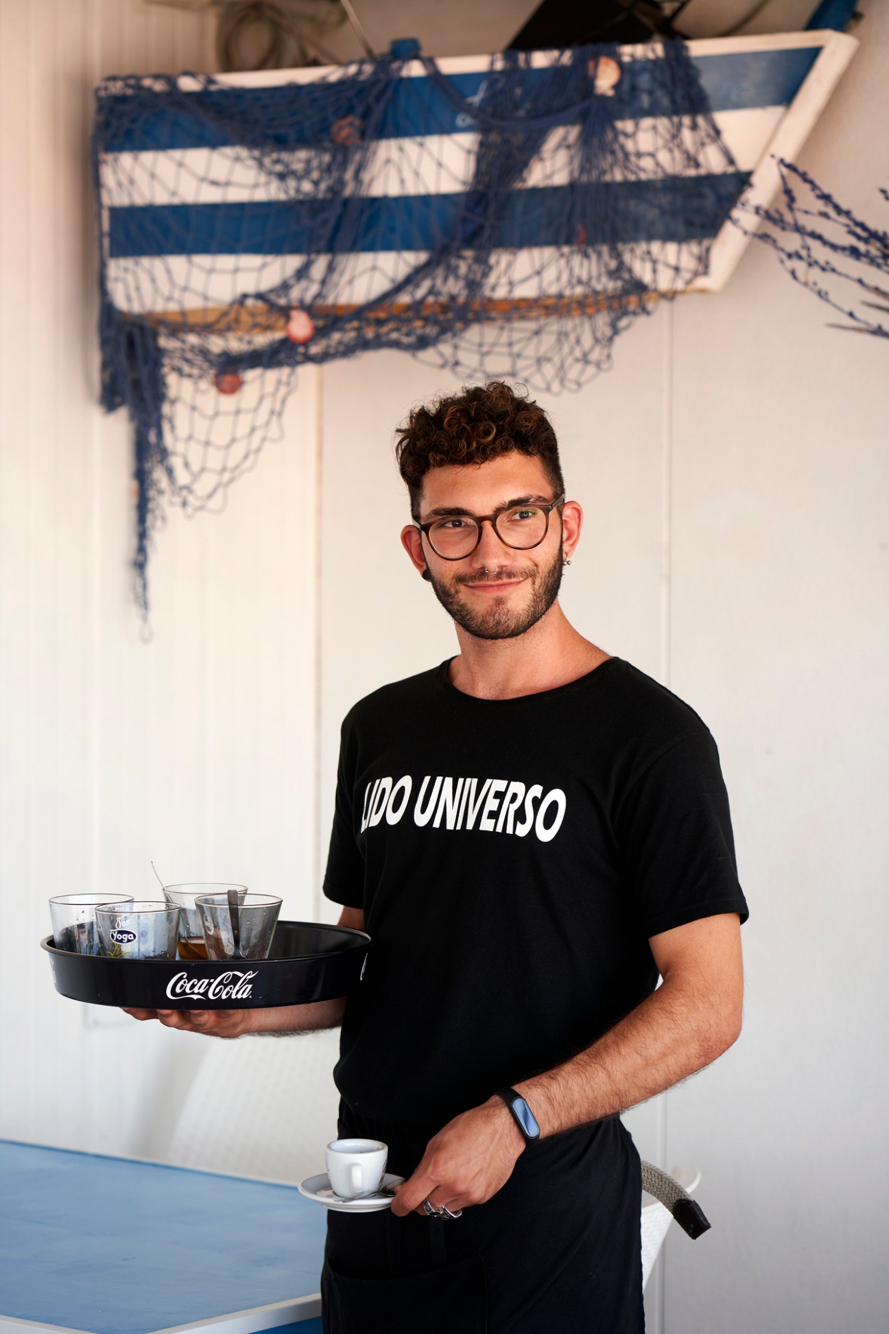 Smiling waiter in black t-shirt carrying drinks tray at Greek seaside restaurant with nautical decor