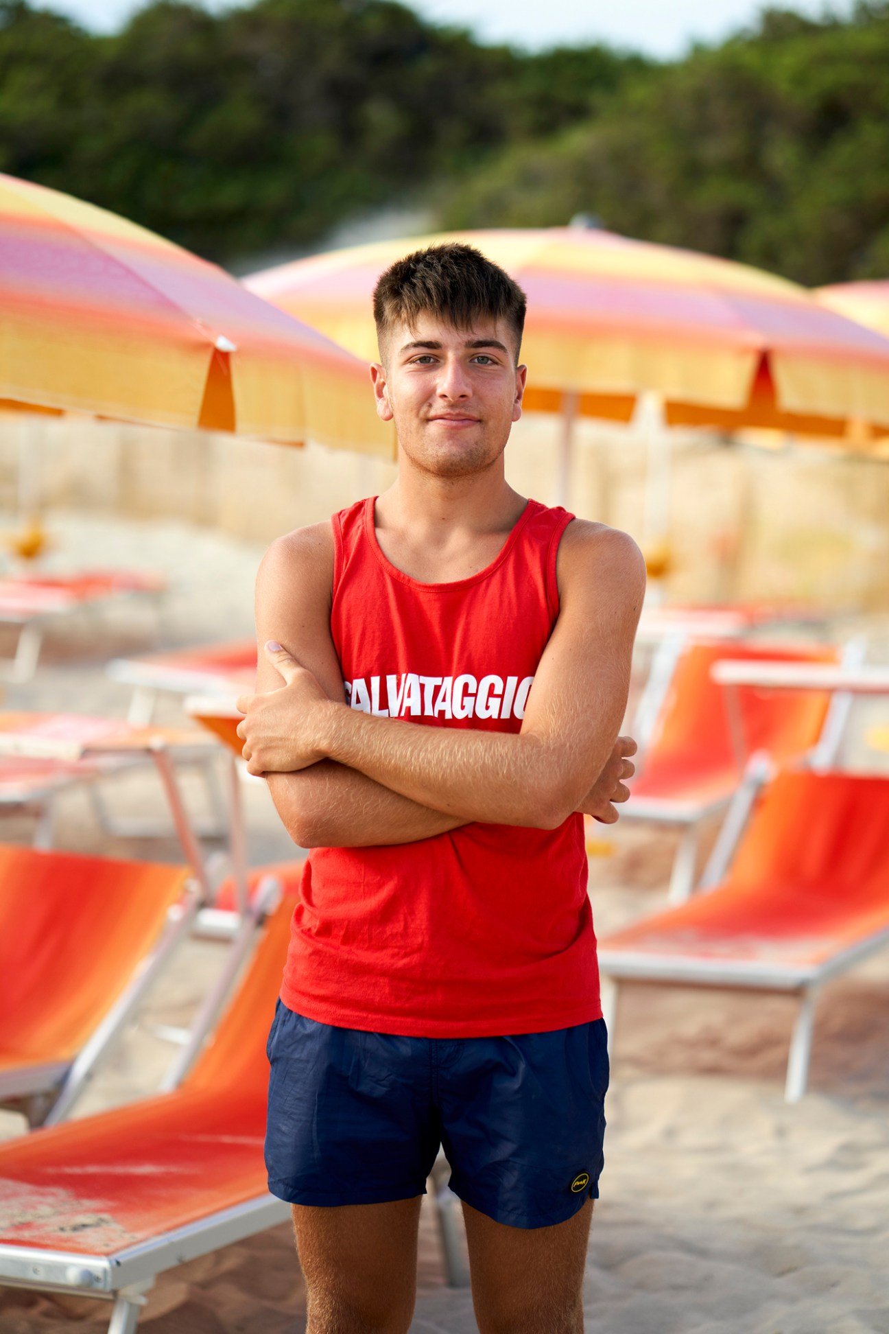 Young man in red tank top with arms crossed standing at Italian beach with umbrellas and lounge chairs