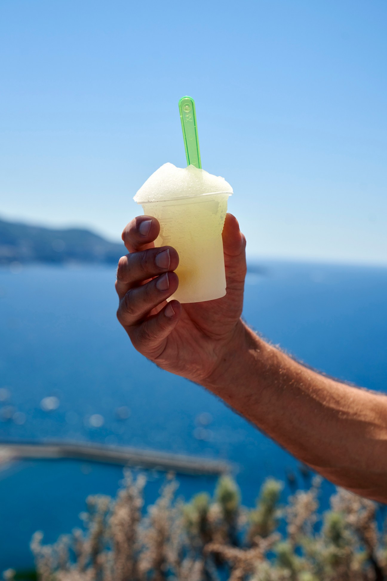 Hand holding frozen drink with green straw against blue sea and sky background
