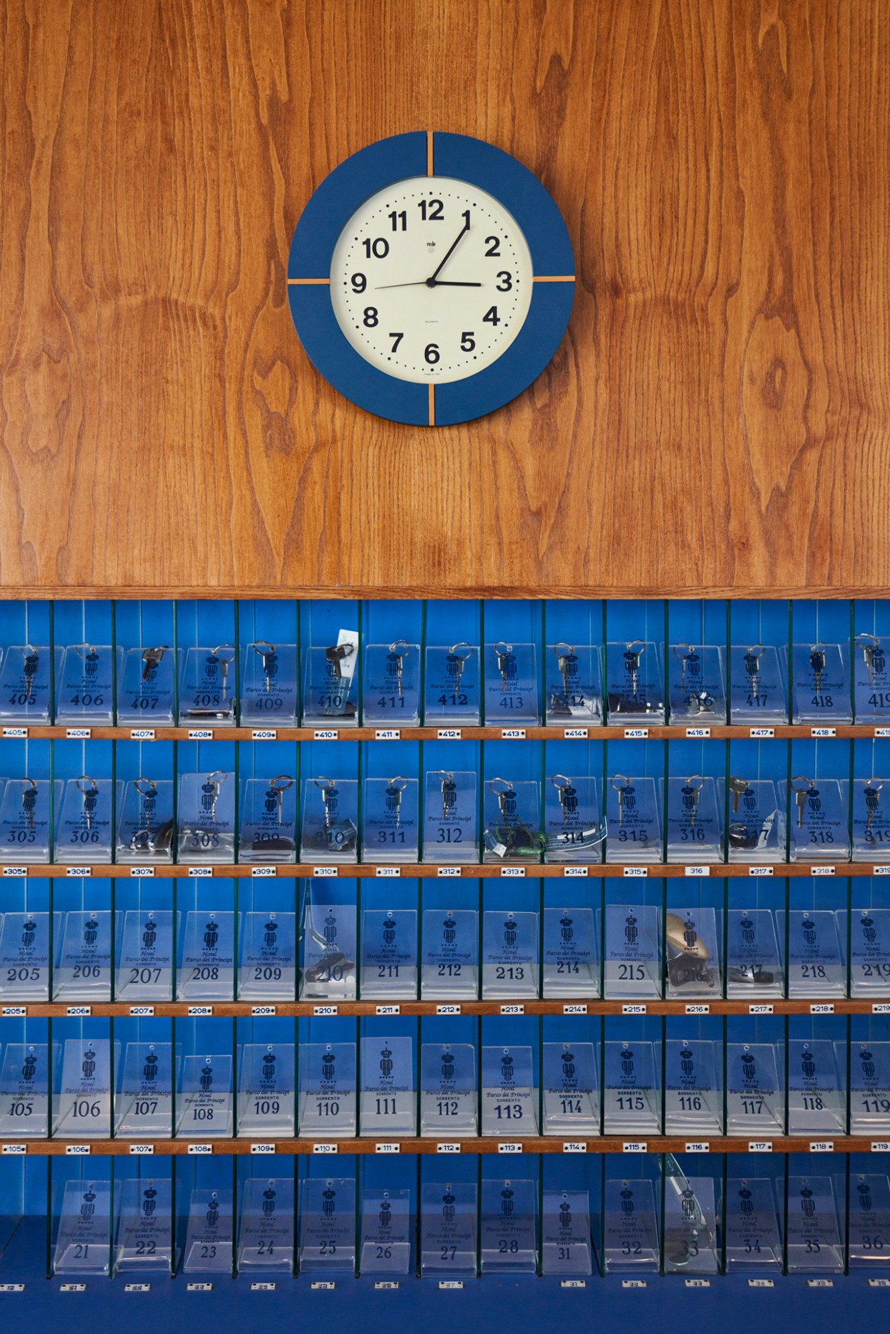 Blue and white hotel room keys hanging in numbered slots from 21 to 419 with a round clock above