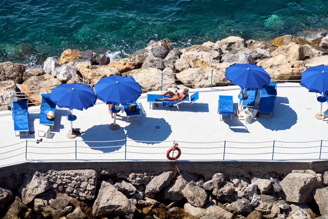 Aerial view of Italian beach club with blue umbrellas, white loungers, and people sunbathing on terrace above rocky coast