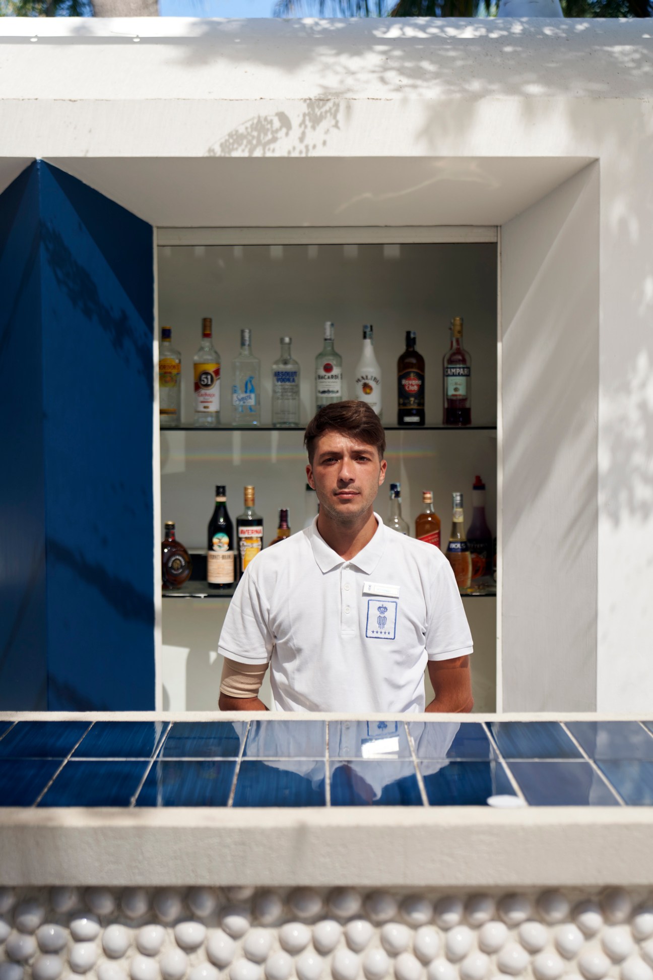Bartender in white polo shirt standing behind modern poolside bar with bottles displayed on shelves