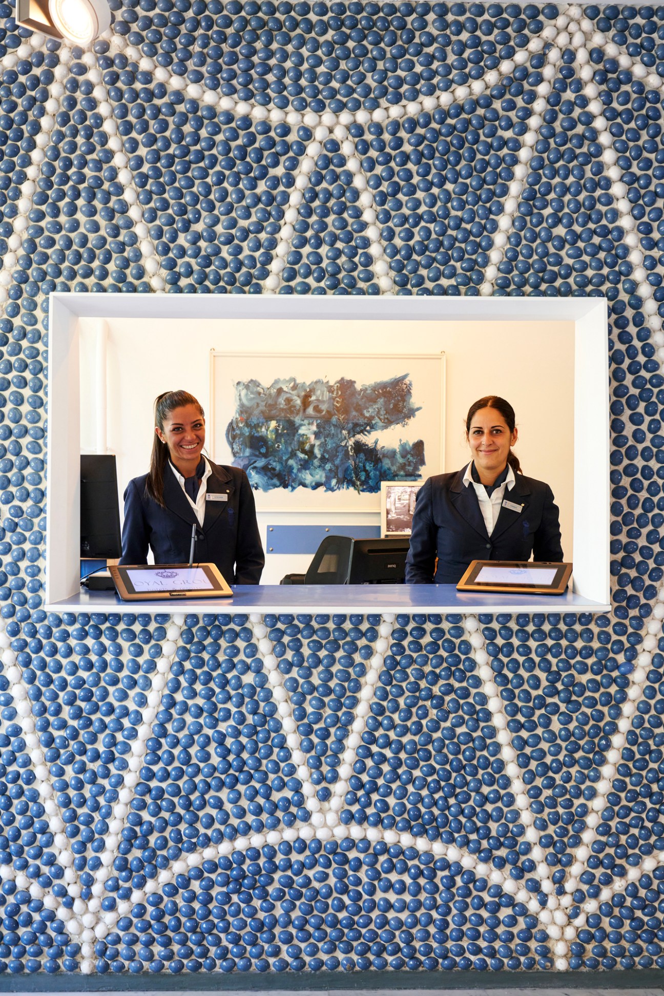 Two female hotel receptionists in navy uniforms standing behind white reception desk with decorative blue ceramic wall
