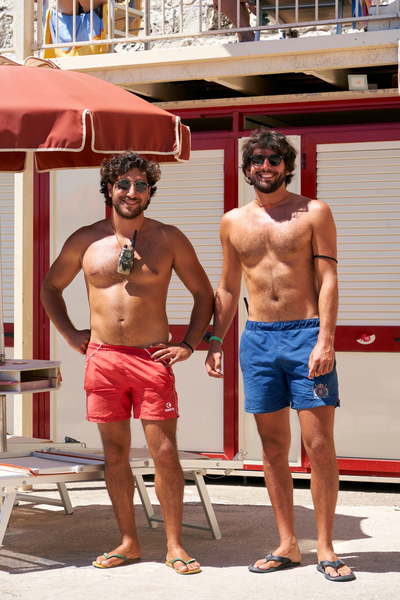 Two shirtless men in colorful swimming shorts standing at an Italian beach establishment