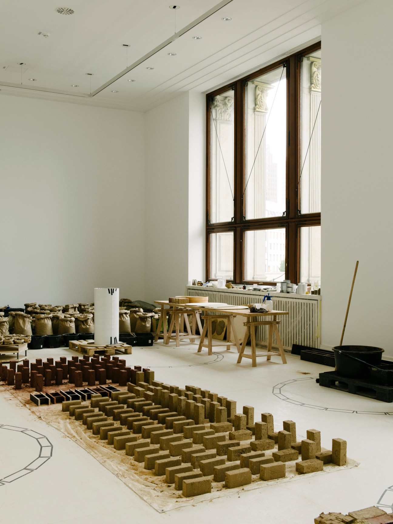 White museum gallery with large window, wooden tables, and floor installation of brown clay blocks and materials