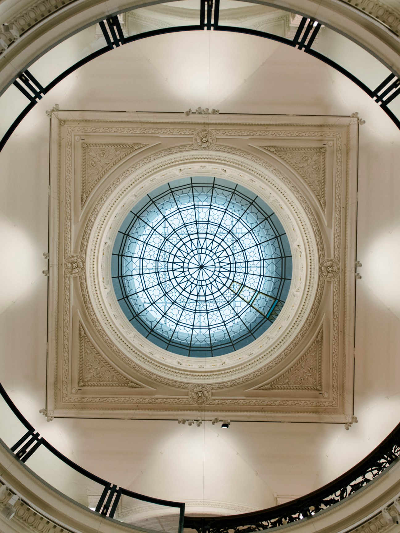 Upward view through circular opening showing ornate ceiling with geometric glass dome and decorative molding