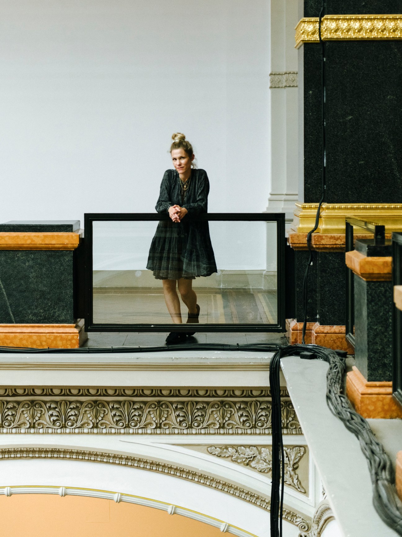 Woman in black dress standing behind glass barrier in ornate museum interior with marble columns