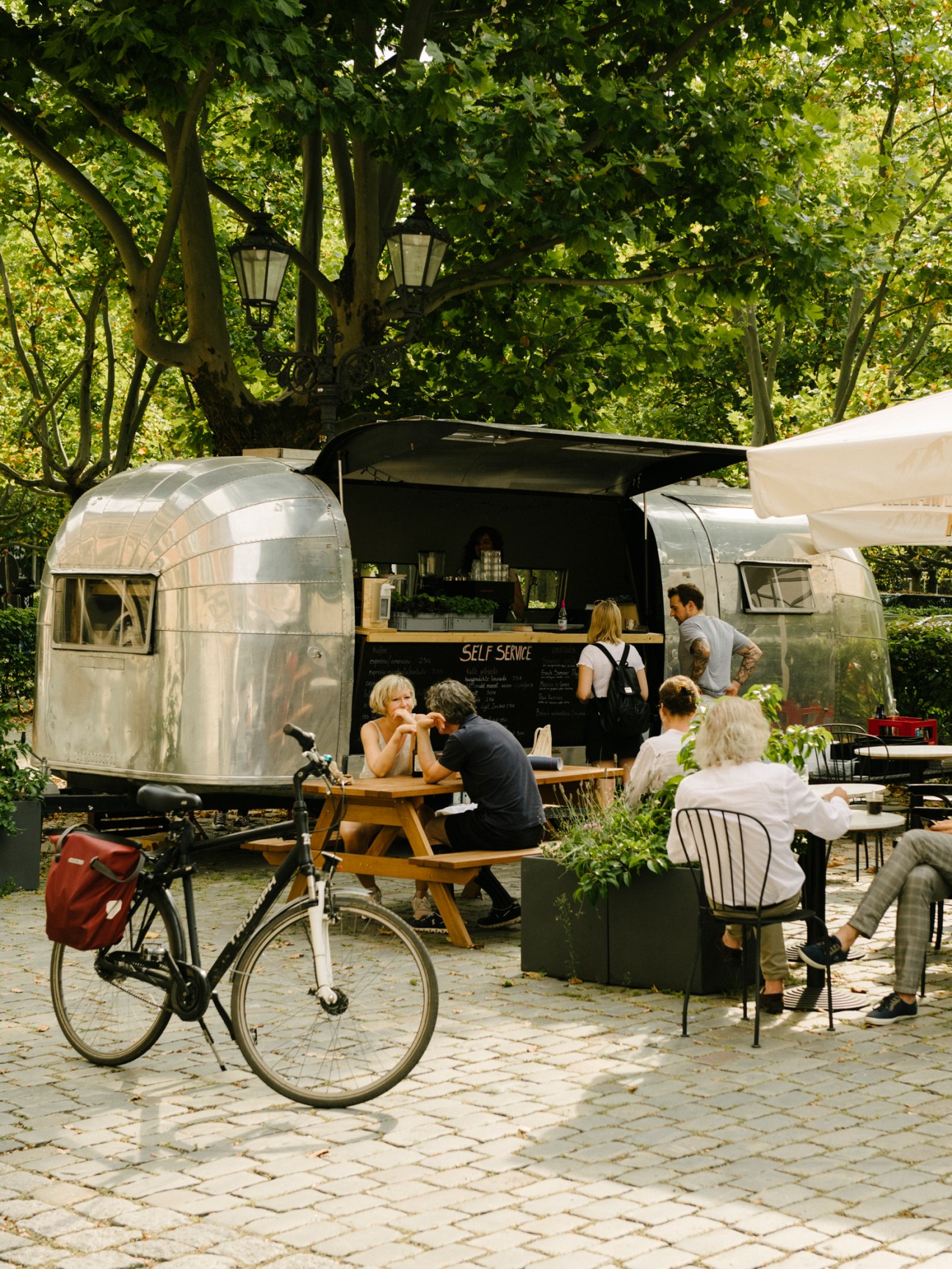 Silver Airstream-style food trailer with service window open in cobblestone courtyard with outdoor seating