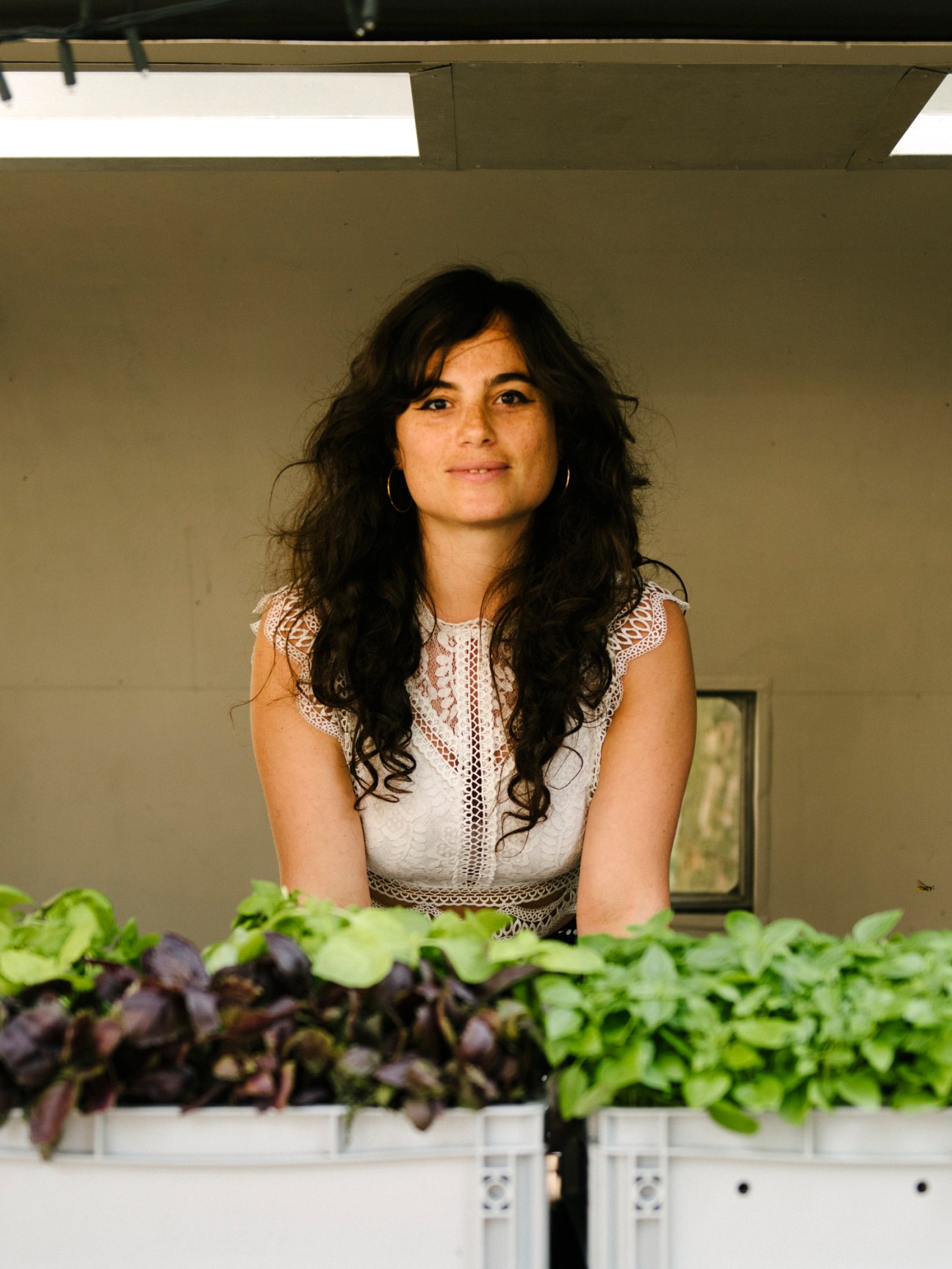 Woman in white lace top with dark wavy hair standing behind leafy green plants in containers