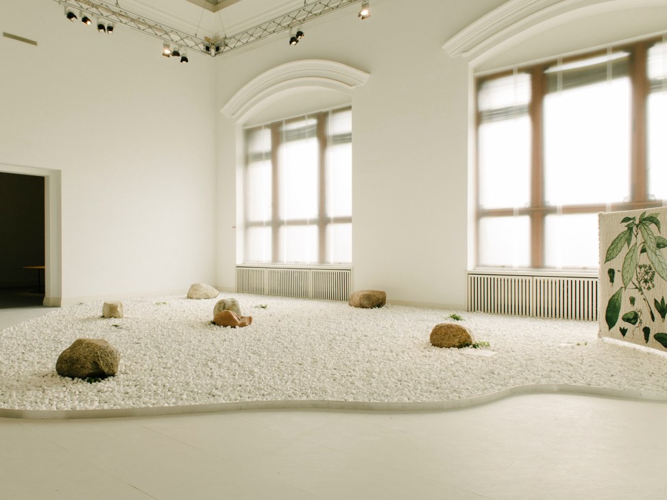 Contemporary art installation with white gravel, brown organic forms, and green plants in museum gallery space