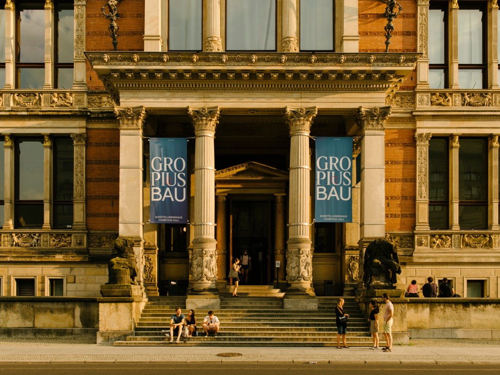 Neoclassical museum building facade with ornate columns, decorative stonework, and blue GROPIUS BAU exhibition banners