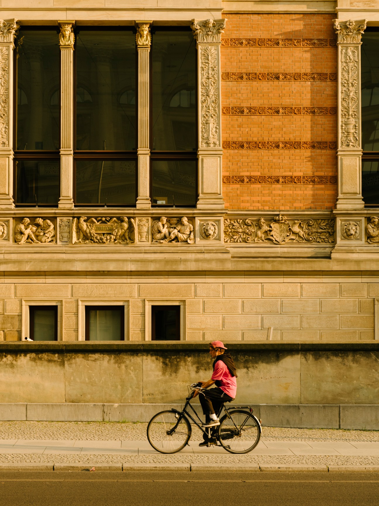 Person in red shirt and cap riding bicycle past ornate Renaissance-style stone building facade