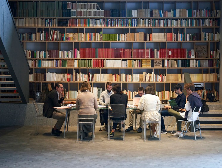 Editorial team of Linkiesta newspaper seated at a table in front of floor-to-ceiling bookshelves in Milan office.
