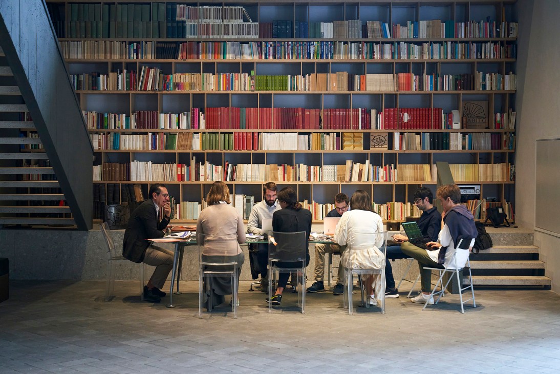 Editorial team of Linkiesta newspaper seated at a table in front of floor-to-ceiling bookshelves in Milan office.