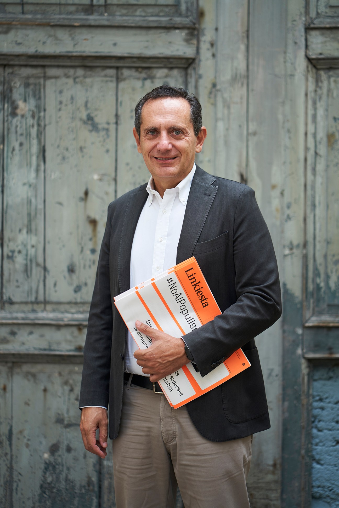 Cristian Rocca, editor of Linkiesta, holds orange and white branded newspapers against a weathered wooden door.