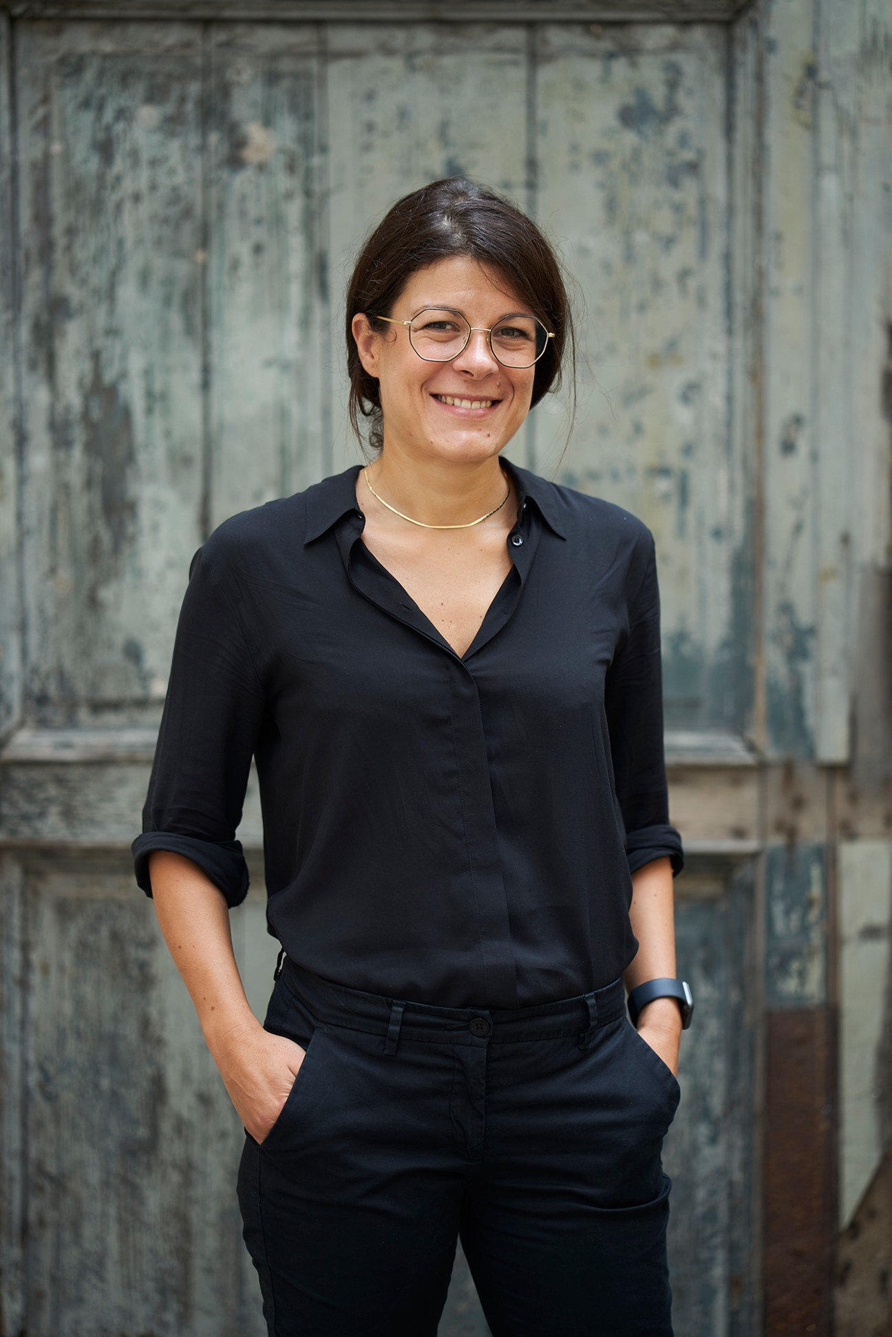 Woman wearing glasses and black shirt smiling at camera against textured wall background.