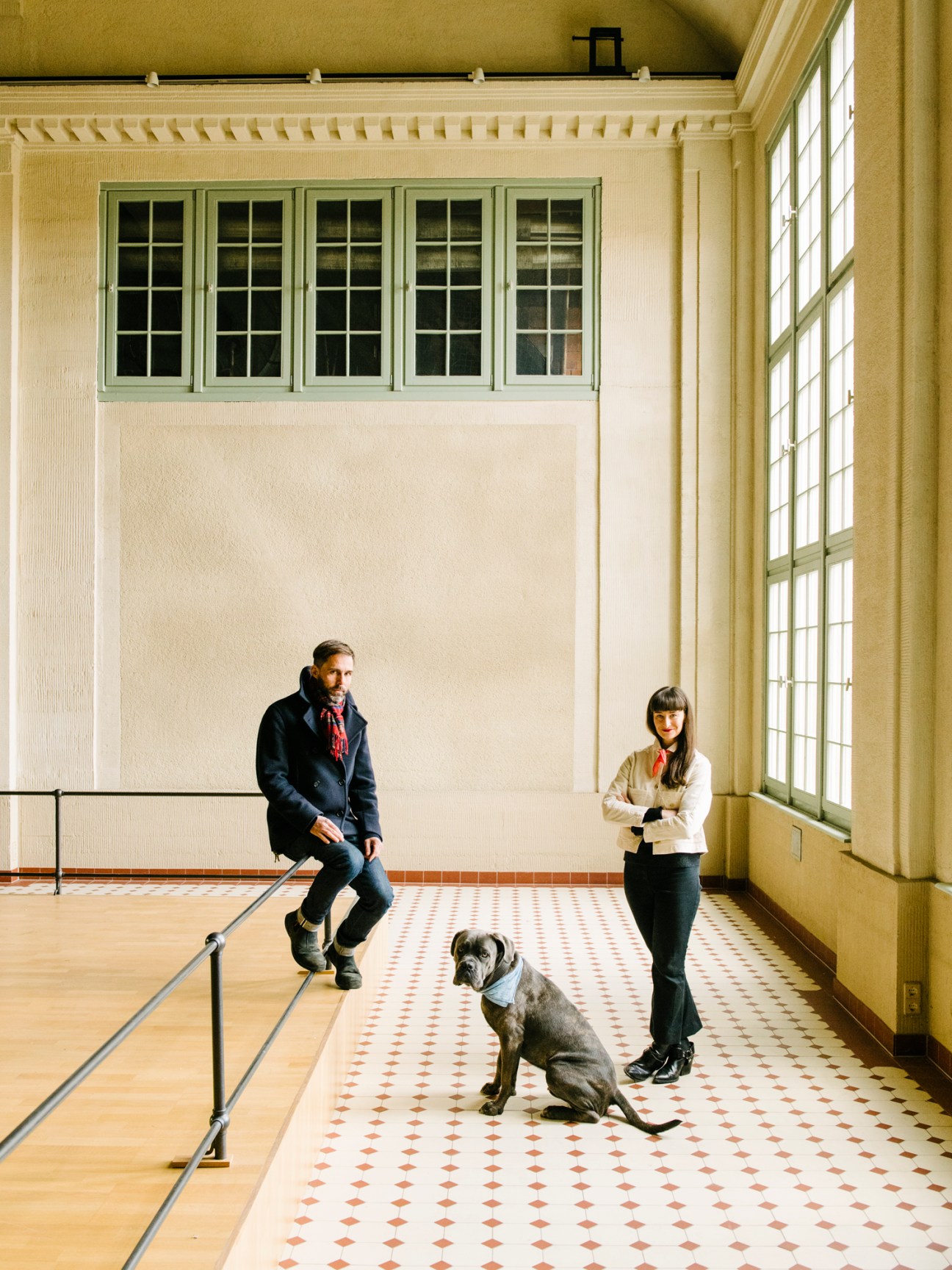 Pablo Wendel, Helen Turner and their dog Coal standing in an industrial space with large windows and tiled floors