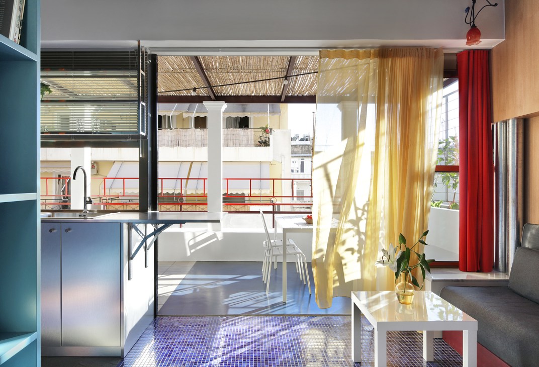 Modern apartment interior with blue kitchen, yellow curtains, white table, and view to terrace with woven shade canopy